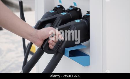 Hand holding EV charging plug in front of camera with blurred background of outdoor. Driver of electric car hold batteries charger plug. Sustainable e Stock Photo