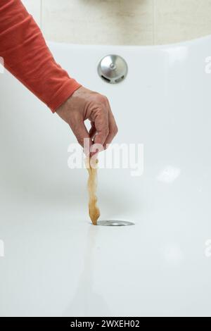 detail of a man's hand, wearing a red t-shirt, pulling a lock of hair out of the drain of a white bathtub. front view. after washing his dog. Hair loss Stock Photo