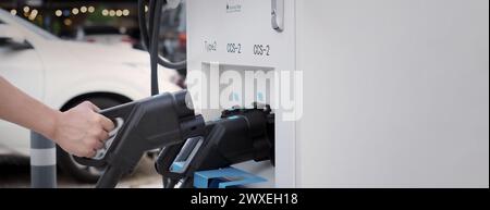 Hand holding EV charging plug in front of camera with blurred background of outdoor. Driver of electric car hold batteries charger plug. Sustainable e Stock Photo