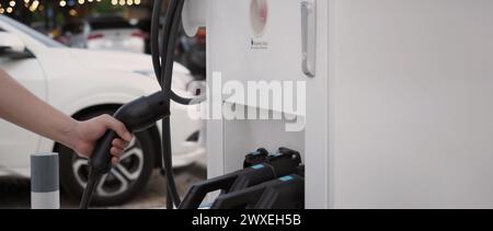 Hand holding EV charging plug in front of camera with blurred background of outdoor. Driver of electric car hold batteries charger plug. Sustainable e Stock Photo
