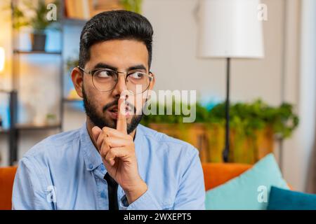 Shh be quiet please. Portrait of young Indian man presses index finger to lips makes silence gesture sign do not tells secret, stop talk gossip, confidential privacy. Guy sitting on couch at home room Stock Photo