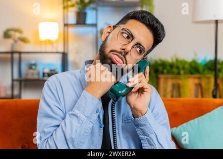 Irritated young Indian man talking on vintage retro wired telephone at home apartment. Disinterested Hispanic guy having annoyed boring conversation with hotline helpline service sitting on sofa. Stock Photo