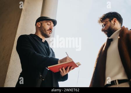 Outdoor business meeting with two stylish male entrepreneurs sharing ideas and writing notes in a red notebook. Stock Photo