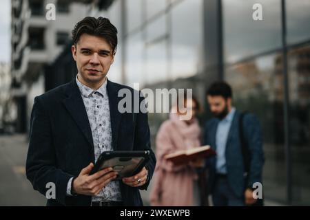 Confident businessman using tablet with colleagues discussing in background outdoors Stock Photo