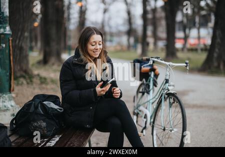 A cheerful young woman sits on a wooden bench in the park, engaging with her smart phone, with a vintage bicycle and backpack beside her. Stock Photo