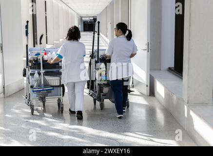 Two female cleaners with cleaning carts walk along a long white corridor. Cleaning concept in office and hospital. Stock Photo