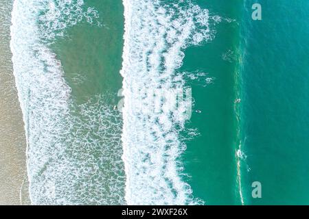 Foam waves breaking near the beach, turquoise sea and two surfers, seen from a drone. Aerial top view Stock Photo