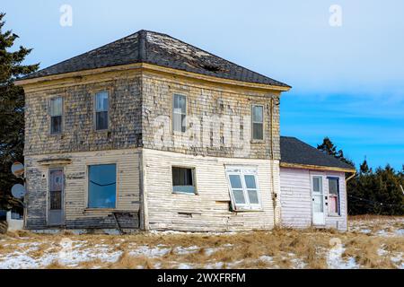 An old abandoned house. The roof has a hole, and a windows is falling out from the side. Rusty satellite dishes on the front, and snow in the grass. Stock Photo