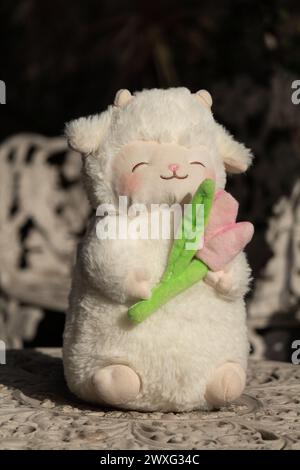 Cute fluffy plush toy of white lamb blushing and holding a pink tulip sitting on top of a white ceramic table Stock Photo