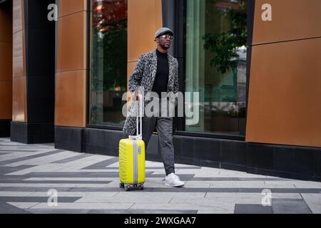 Stylishly dressed male model walks down city street with yellow suitcase in hand. Black man tourist outdoors near hotel building. African american man Stock Photo