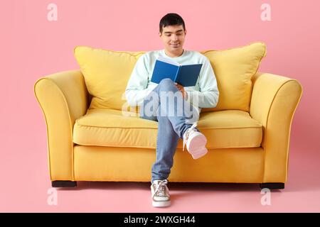 Handsome young man reading book on yellow sofa against pink background Stock Photo