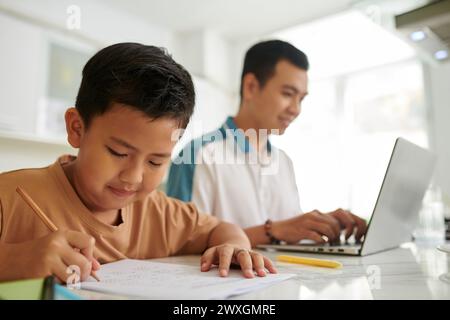Boy doing homework when sitting next to father working on laptop at kitchen counter Stock Photo