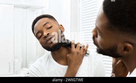 Man trimming beard with electric clippers in mirror in bathroom Stock Photo