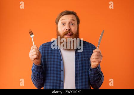 Hungry redhaired bearded guy holding knife and fork, orange background Stock Photo