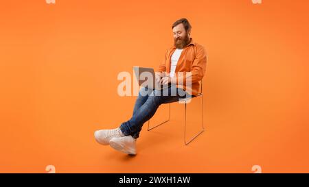 redhaired programmer guy sits typing on his laptop, orange background Stock Photo
