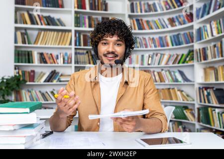 Cheerful young male educator engaging in online teaching using a headset and video call technology in a library setting. Stock Photo