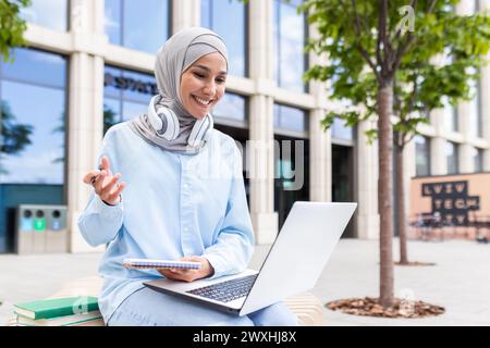 Focused young student wearing a hijab with headphones studying on her laptop and taking notes outside on campus. Stock Photo
