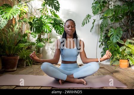 Calm african american girl in lotus pose meditates in space filled with tropic plants stress relieve Stock Photo