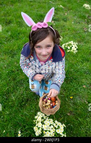 Easter egg hunt smiling girl child 6 7 wearing rabbit ears holding basket of chocolate eggs  in  Wales March garden 2024 UK Great Britain KATHY DEWITT Stock Photo