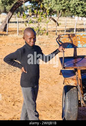 smiling african child standing next to a rusty donkey cart in the village at sunset, Stock Photo