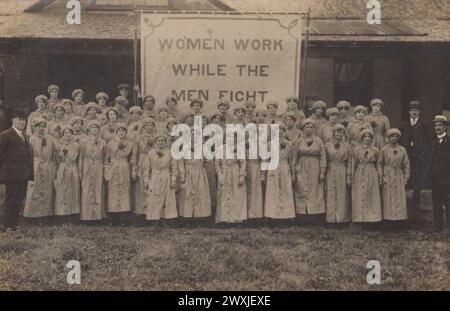 'Women Work While The Men Fight': photograph of female munitions workers outside the Siemens factory in Stafford during the First World War (probably 1916 or 1917). The women, and the banner that they're standing in front of, took part during the annual hospital pageant or parade. The women munitions workers are all dressed in their working clothes - long overalls and close fitting caps - and have a sprig of flowers or foliage attached to their lapels Stock Photo