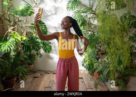 Happy joyful black woman smiling makes selfie on smartphone stands in room with tropical houseplants Stock Photo