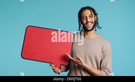 Happy man holding red speech bubble sign of empty space for text. Cheerful Middle Eastern person presenting thought bubble cardboard, isolated over blue studio background Stock Photo