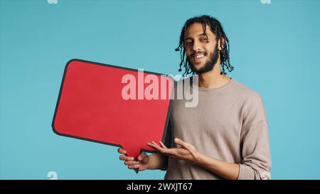 Smiling BIPOC man holding red speech bubble sign of empty copy space for message. Joyous person presenting thought bubble cardboard used as promotion concept, isolated over studio background Stock Photo