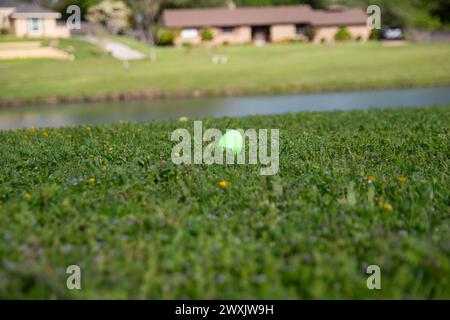 A green, plastic Easter egg hidden in the short grass in a neighborhood park with a pond for a holiday celebration. Stock Photo