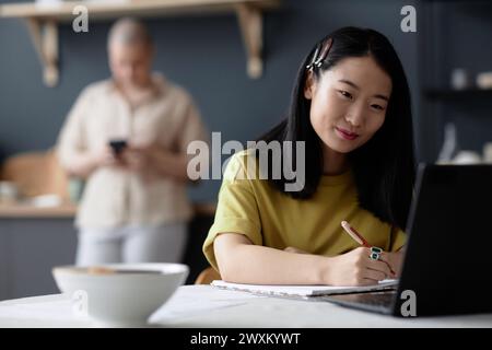 Young female Asian student sitting at dining table looking at laptop screen and making note in notebook Stock Photo
