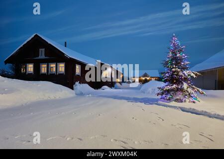 Christmas Eve in village in Northern Europe, Christmas tree stands on street near wooden house in evening. Stock Photo