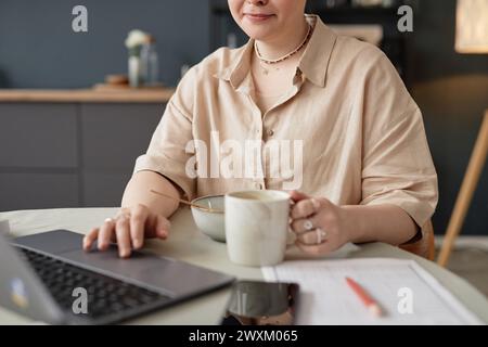 Medium crop shot of young Caucasian woman sitting at table in kitchen having breakfast and scrolling web page in Internet on laptop Stock Photo
