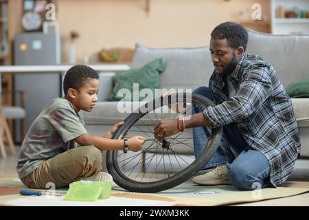 Side view portrait of African American father and son repairing bicycle wheel together sitting on floor at home Stock Photo