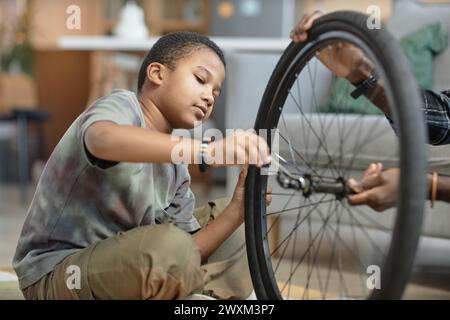Side view portrait of young Black boy repairing bicycle wheel with father sitting on floor at home Stock Photo