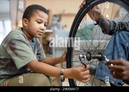 Side view portrait of young African American boy repairing bicycle tire with dad sitting on floor at home Stock Photo
