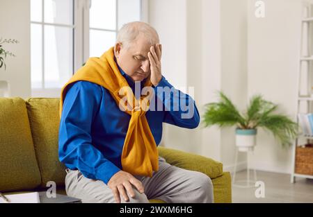 Portrait of gray-haired senior man suffering from headache disease sitting on sofa at home. Stock Photo