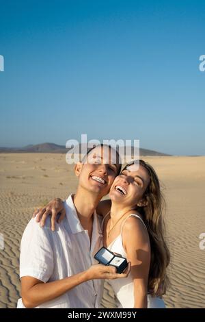Happy young couple with a proposal ring box, celebrating engagement in a scenic desert Stock Photo