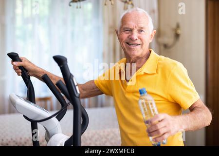 Senior man with bottle of water using elliptical trainer at home Stock Photo