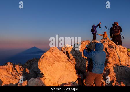 Hikers standing on the summit of Mount Teide at sunrise enjoying panoramic views and taking photos, with the mountain cone shadow on the horizon Stock Photo
