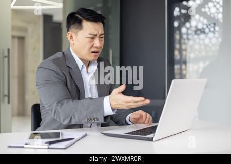 Asian businessman in grey suit looking confused while working on his laptop at a modern office workspace. Stock Photo