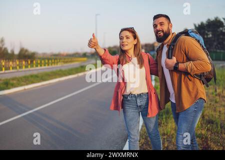 Happy couple hitchhiking on roadside trying to stop car. Stock Photo