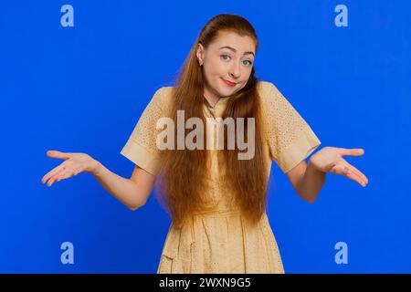 What Why. Sincere irritated young woman raising hands in indignant expression, ask reason of failure demonstrating disbelief irritation by trouble. Confused redhead girl on blue background, indoors Stock Photo