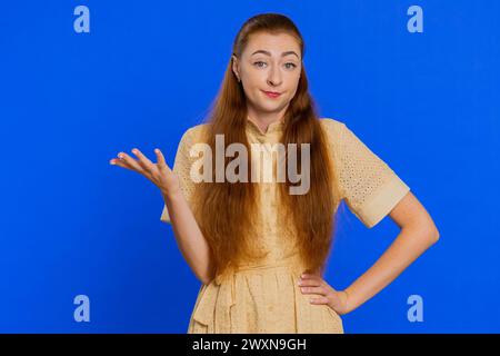 What Why. Sincere irritated young woman raising hands in indignant expression, ask reason of failure demonstrating disbelief irritation by trouble. Confused redhead girl on blue background, indoors Stock Photo