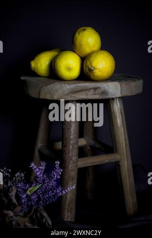 A still life with lemons and purple flowers on a stool against a dark backdrop Stock Photo