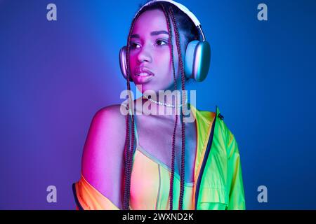 pensive dark-skinned woman in wireless headphones posing in jacket in studio with neon lights Stock Photo