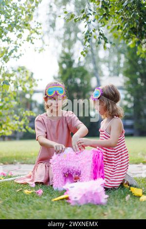 Two girls smashing and opening pink pinata with a stick at birthday party. Children celebrating birthday at garden party. Children having fun and play Stock Photo