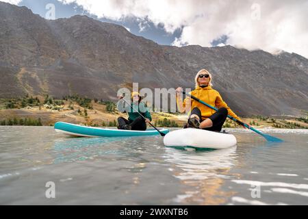 Two surfers girls sits and walks on sup boards at calm mountain lake with white water Stock Photo