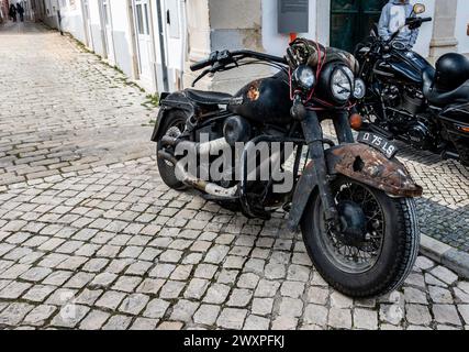 A rusty, vintage motorcycle with distinct patina stands parked on an old cobblestone street, in Loule, Portugal. Stock Photo