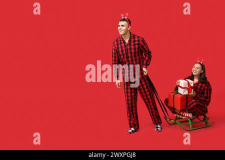 Happy young couple in Christmas pajamas, with sledge and gifts on red background Stock Photo