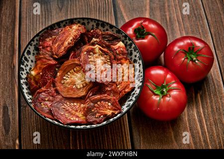 Sun dried tomatoes in bowl and three fresh ripe tomatoes on dark wooden table background. Top view. Stock Photo